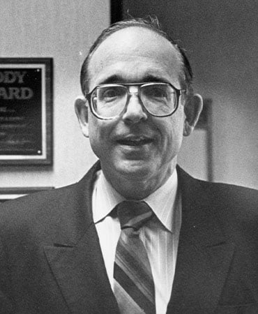 Black and white photo of Lawrence Kugler wearing a suit, dress shirt, and striped tie, standing indoors with a framed certificate on the wall behind him.