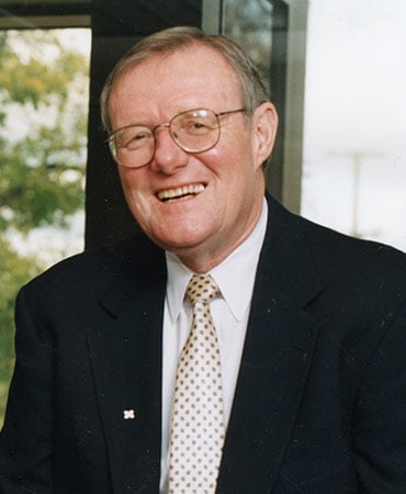 Marvin Roberson wearing a black suit jacket, white shirt, and patterned tie standing indoors near a window with trees visible outside.