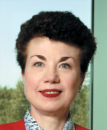 Ruth Person with short curly black hair wearing a red jacket and a patterned shirt standing indoors near a window with trees visible outside.