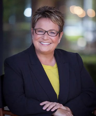 Susan Borrego wearing a dark blazer over a yellow top, sitting with hands crossed, with a blurred background of lights and greenery.