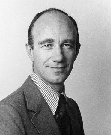 A black and white portrait of William Moran wearing a suit and a striped shirt, with a blurred face.