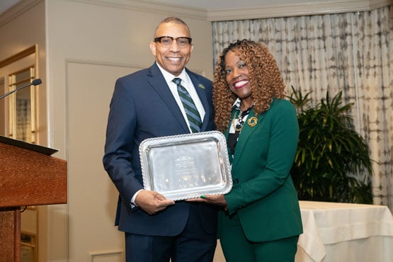 Two people in formal attire holding a silver award plaque together in an indoor setting.