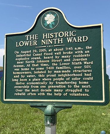 Green historical marker sign titled “The Historic Lower Ninth Ward” with text describing the August 29, 2005, Industrial Canal flood wall break during Hurricane Katrina, its impact on residents, and the neighborhood’s struggle to rebuild. A colorful mural is visible on a wall to the left, with a residential street and houses to the right under a clear blue sky.