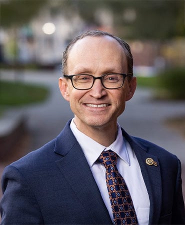 Chris Lewis wearing a dark blue suit, white shirt, and patterned tie standing outdoors on a pathway with greenery in the background.