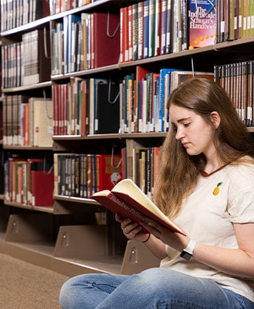 Person with long hair sitting on the floor in a library, reading a book with bookshelves in the background.