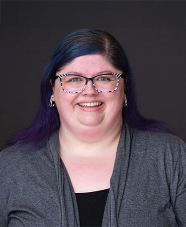 Kim Saks with long dark hair wearing gold hoop earrings, a gray cardigan, and a black top against a dark background.