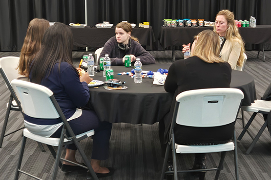 Five women sit at a black-clothed round table, engaged in conversation. The table is covered with snack wrappers, bottled water, and soda cans. The setting includes folding chairs, a dark curtain backdrop, and tables in the background stacked with more snacks and drinks.