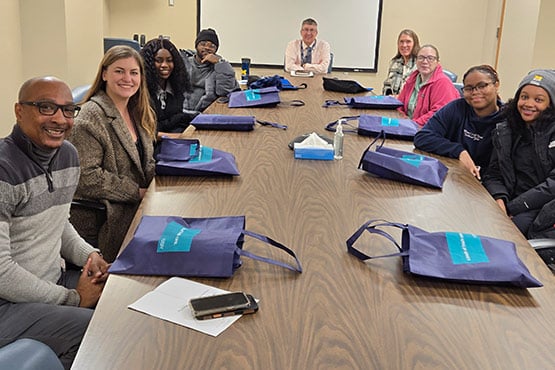 A group of people sitting around a conference table with blue bags in front of them and a presentation screen in the background.