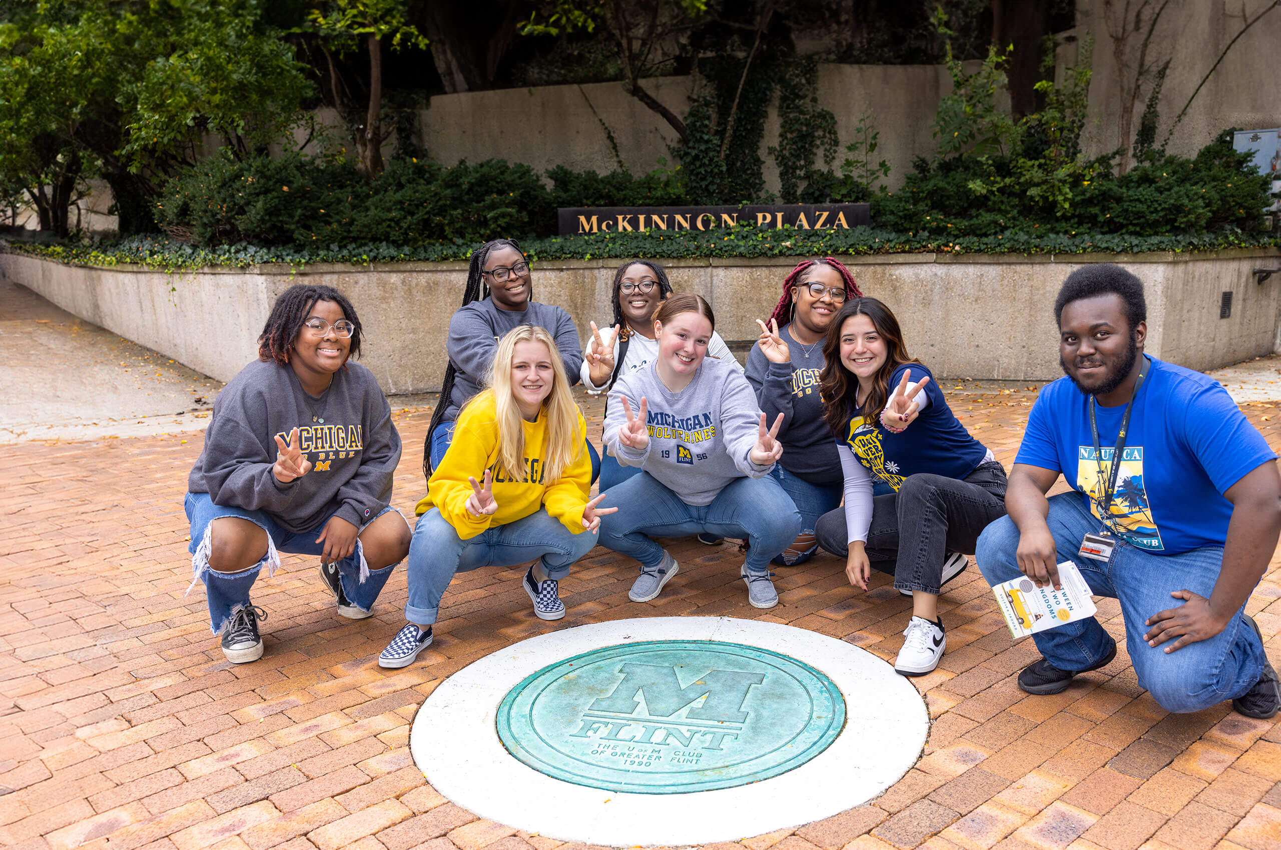 A group of seven people poses in front of a circular emblem on the ground that reads "M FLINT" in McKinnon Plaza. They are wearing sweatshirts and making peace signs.