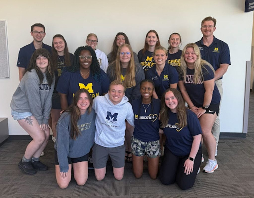 A group photo of 15 individuals, mostly wearing apparel from the University of Michigan, posing together in a well-lit indoor space.