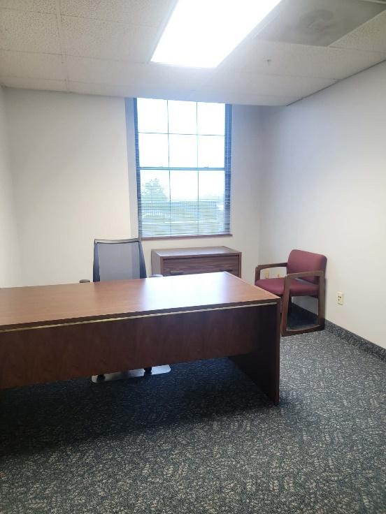 Office room with a wooden desk, a mesh office chair, a small wooden cabinet under a window with blinds, and a red guest chair against a white wall.