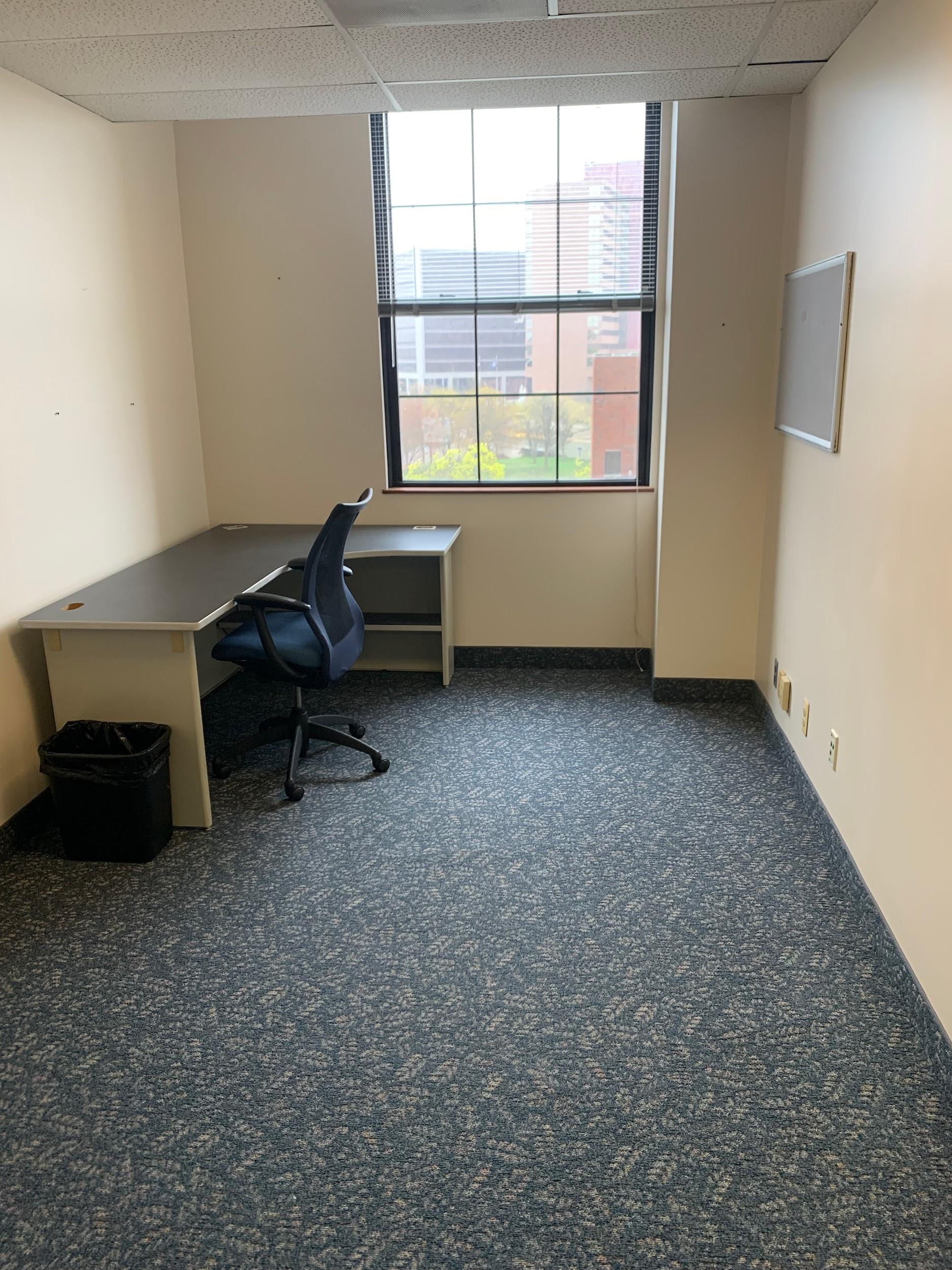 Small empty office with a desk, office chair, trash bin, window with city view, and a whiteboard on the wall.