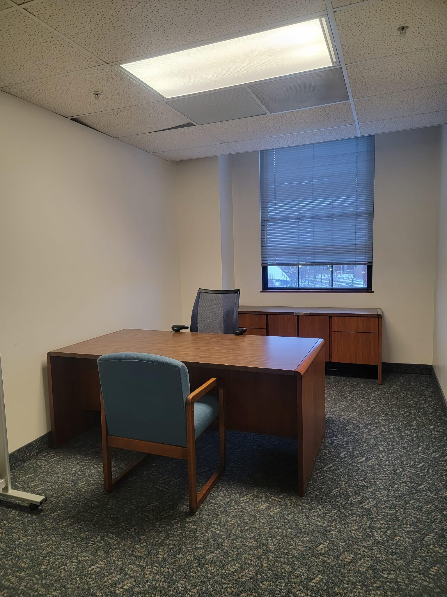 Small office with a wooden desk, two chairs, a window with blinds, and a wooden cabinet against the wall.