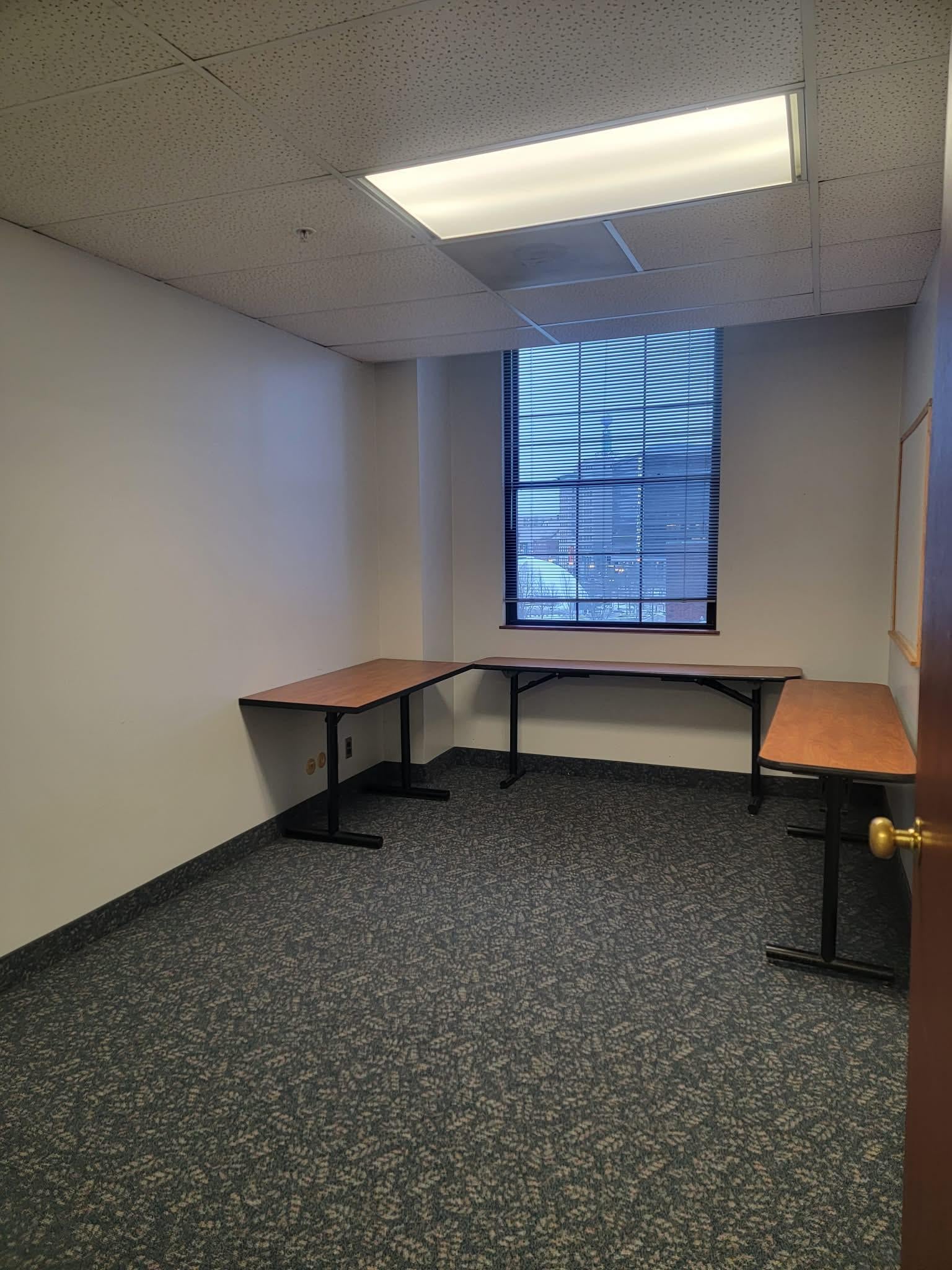 Empty office room with three wooden tables arranged in a U-shape under a window with blinds, carpeted floor, and fluorescent ceiling light.