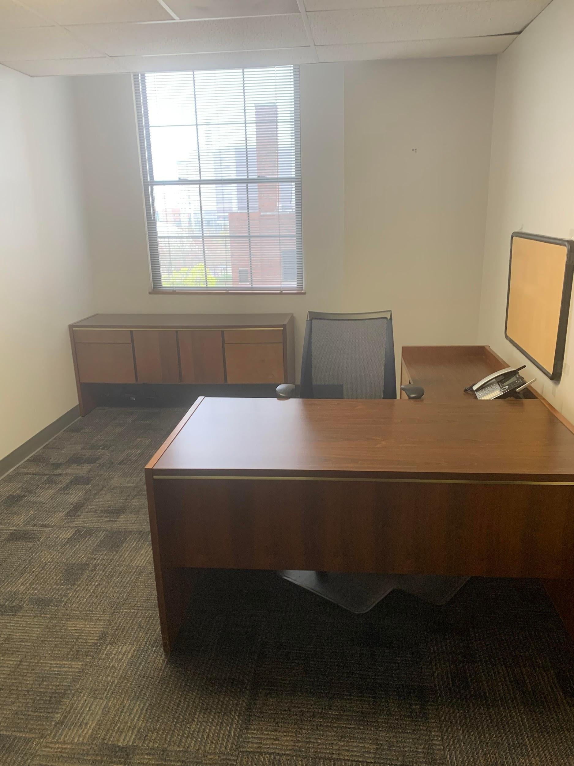 Small office with a wooden desk, office chair, side cabinet, window with blinds, and a phone on the desk.
